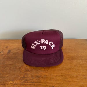 Op de afbeelding: Bordeaux truckerpet met een mesh achterkant en een gebogen klep. Het voorpaneel toont de witte tekst "SIX-PACK 19" in een gebogen, gestileerd lettertype. De pet ligt op een houten oppervlak.