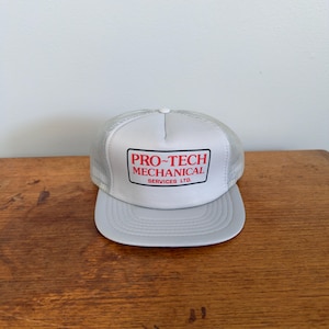 以下が含まれることがあります： 前面に赤と黒の長方形のパッチで「PRO-TECH MECHANICAL SERVICES LTD.」の文字が入った、グレーと白のトラッカーハット。帽子にはグレーのつばと白いメッシュの背面が付いています。
