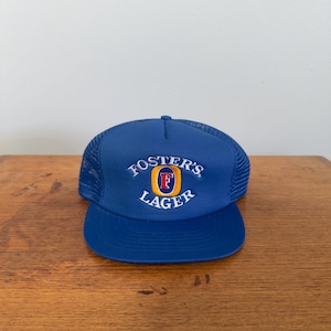 Op de afbeelding: Een blauwe truckerpet met een gebogen klep en mesh achterkant. De pet heeft het Foster's Lager-logo in wit, rood en geel op de voorkant geborduurd. De pet ligt op een houten oppervlak.