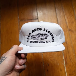 Op de afbeelding: Witte truckerpet met een zwart-witte afbeelding van een auto en de tekst "STAR AUTO ELECTRIC DOWNSVIEW ONT."