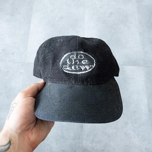 Puede incluir: Gorra de béisbol negra con visera curva. La gorra presenta el texto "do the dew" en un parche ovalado blanco en la parte delantera. El sombrero está hecho de tela oscura.