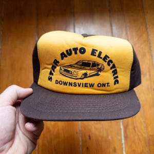 Op de afbeelding: Bruine en gele truckerpet met de tekst "STAR AUTO ELECTRIC" en een autografiek. De pet heeft ook de tekst "DOWNSVIEW ONT." op de voorkant. De pet heeft een bruine klep en een geel voorpaneel.