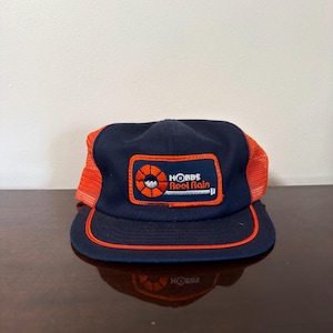 Peut inclure: Une casquette de baseball bleu marine avec des panneaux latéraux en maille orange et un bord orange. L'avant présente un écusson rectangulaire avec un logo circulaire orange et le texte Hobbs Reel Rain en blanc et orange sur fond bleu.