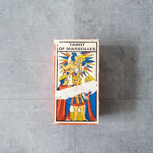 May include: A rectangular box of Tarot cards, with the text "TAROT OF MARSEILLES". The box has a colourful illustration of figures and an angel. The box is orange, red, blue, yellow, and white.