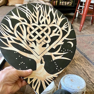 May include: A circular wooden plaque featuring a black painted background with a natural wood grain tree design. The tree branches form a central heart shape with an intricate Celtic knot pattern inside. Small white dots are scattered around the base.