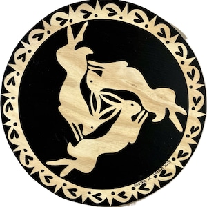 May include: A black circular wooden plaque with a white decorative border. Three stylised white rabbits are depicted in a circular pattern on the black background. The plaque is signed "Hunter M. Yoder 2024".