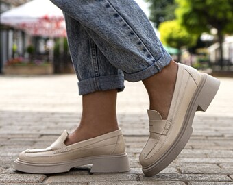 May include: A pair of light beige leather loafers with a chunky platform sole. The loafers have a simple design with a single strap across the top and a small buckle detail.