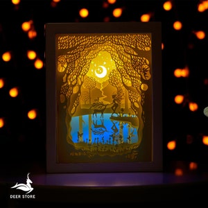 Puede incluir: Una lámpara de papel recortado blanca con un hada y un ciervo en una escena de bosque. La lámpara está iluminada con una luz amarilla cálida, creando una atmósfera mágica. La escena incluye una luna creciente y estrellas.