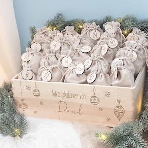 Könnte beinhalten: Ein Adventskalender aus Holz mit kleinen beigefarbenen Kordelzugbeuteln, die jeweils mit einer Zahl von 1 bis 24 beschriftet sind. Die Box ist mit gravierten Weihnachtsschmuck und dem Text "Adventskalender von Paul" verziert.