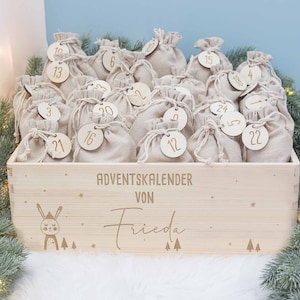 Può includere: Un calendario dell'Avvento in legno con 24 piccole sacche beige con coulisse, ciascuna con un'etichetta di legno numerata da 1 a 24. La scatola del calendario è incisa con le parole "ADVENTSKALENDER VON Frieda", un coniglietto, alberi e stelle.