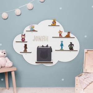 Peut inclure: Étagère murale en forme de nuage blanc avec plusieurs étagères pour exposer des jouets et un petit haut-parleur. L'étagère porte le nom "JONAH" écrit en lettres beiges.
