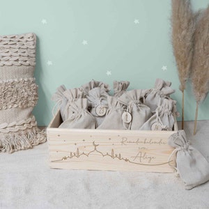 May include: A wooden box with a Ramadan calendar design. The box is filled with small burlap bags, each with a number on it. The box is decorated with a mosque silhouette and stars. The text "Ramadan calendar of Aliya" is engraved on the box.