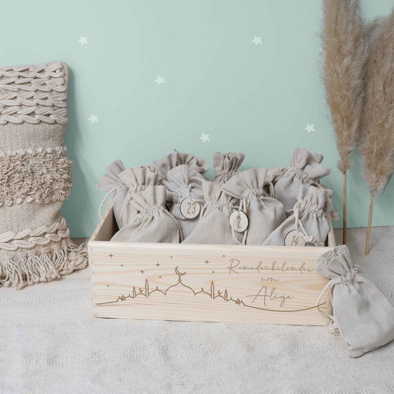 May include: A wooden box with a Ramadan calendar design. The box is filled with small burlap bags, each with a number on it. The box is decorated with a mosque silhouette and stars. The text "Ramadan calendar of Aliya" is engraved on the box.