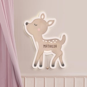Könnte beinhalten: Eine hellbraune, rehförmige Wandleuchte mit dem Aufdruck "MATHILDA". Das Reh hat eine weiße Kontur, eine rosa Wange und einen kleinen Schwanz. Das Licht hängt an einer hellrosa Wand neben einem rosa Vorhang.
