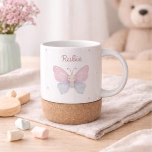 May include: White ceramic mug featuring a pink and blue butterfly design with the name "Rubie" printed above. The mug sits on a cork base. A handle is attached to the side. The mug is decorated with small dots.