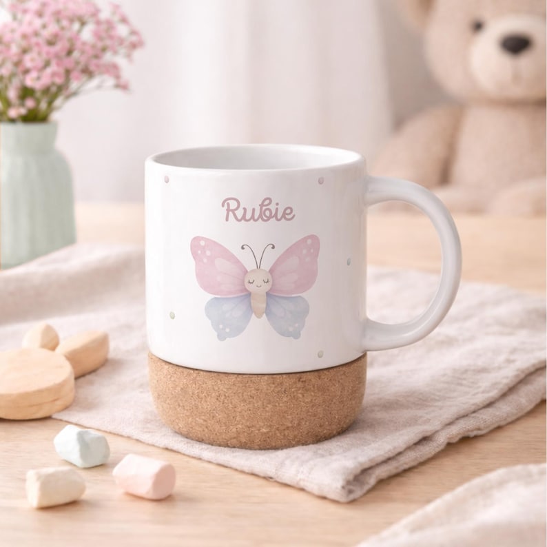 May include: White ceramic mug featuring a pink and blue butterfly design with the name "Rubie" printed above. The mug sits on a cork base. A handle is attached to the side. The mug is decorated with small dots.