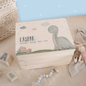 Könnte beinhalten: Eine hellfarbene Holzkiste mit einer Dinosaurier-Illustration und dem Namen "Caspar". Die Kiste zeigt einen grünen und grauen Dinosaurier, Wolken und den Text "05.09.2022 - 14:03 UHR - 3140 G - 52 CM". Die Kiste ist im Baby-Thema dekoriert.