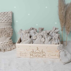 May include: A wooden box with a Ramadan calendar design. The box is filled with small burlap bags, each with a number on it. The box is decorated with a mosque and stars. The text "Ramadan Kalender" is engraved on the box. The box is on a white surface.