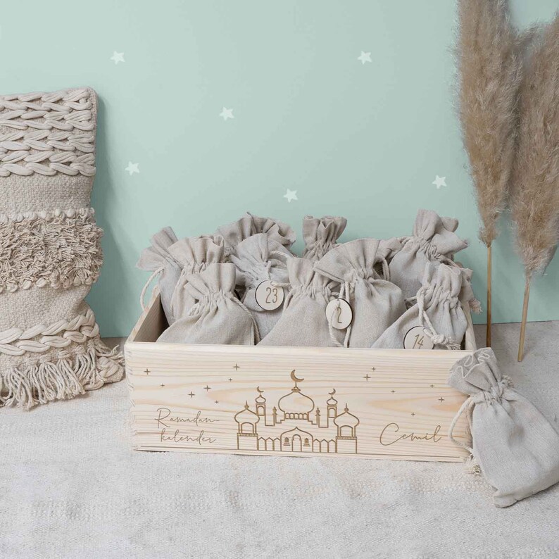 May include: A wooden box with a Ramadan calendar design. The box is filled with small burlap bags, each with a number on it. The box is decorated with a mosque and stars. The text "Ramadan Kalender" is engraved on the box. The box is on a white surface.