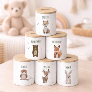 Può includere: Salvadanai in ceramica bianca con coperchi in legno, ognuno con un'illustrazione di un animale del bosco e un nome. Gli animali includono una volpe, un orso, uno scoiattolo, un gufo, un cervo e un coniglio. I nomi sono in verde.
