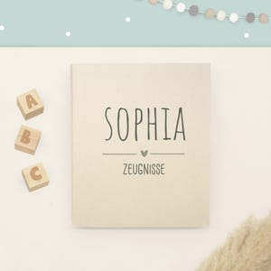 May include: A beige rectangular book with the name SOPHIA printed in dark green capital letters, followed by a small heart icon and the word ZEUGNISSE. Three wooden blocks with the letters A, B, and C are positioned to the left of the book.