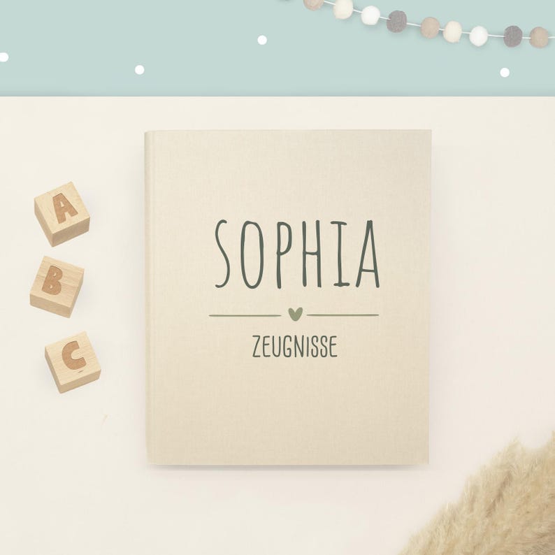 May include: A beige rectangular book with the name SOPHIA printed in dark green capital letters, followed by a small heart icon and the word ZEUGNISSE. Three wooden blocks with the letters A, B, and C are positioned to the left of the book.