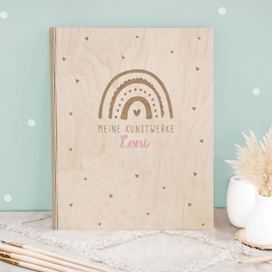 Op de afbeelding: Een houten doos met een regenboogontwerp en de tekst "Meine Kunstwerke Leni" in roze letters. De doos is versierd met kleine hartjes.