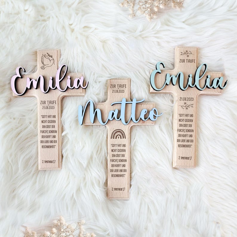 May include: Three wooden crosses with the names Emilia, Matteo, and Emilia, each with a different color scheme. The crosses are decorated with a dove, a rainbow, and floral designs. The crosses also include the text "Zur Taufe" (German for "for baptism") and the date "21.08.2023".
