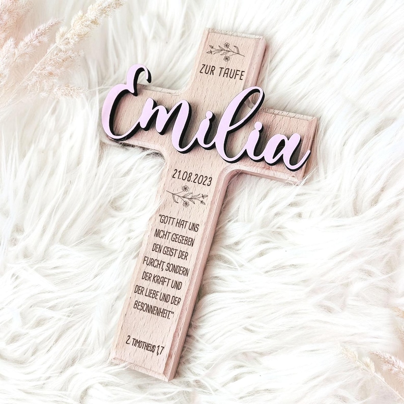May include: A wooden cross with the name "Emilia" in pink lettering, the date "21.08.2023", and the text "Zur Taufe" (German for "For Baptism"). The cross also features a Bible verse from 2 Timothy 1:7.