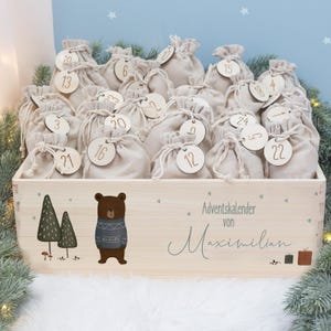 Op de afbeelding: Een houten adventskalenderbox gevuld met kleine beige stoffen zakjes, elk met een houten label met nummer 1-24. De doos is versierd met een beer met een trui en twee gestileerde bomen. De tekst "Adventskalender von Maximilian" staat ook op de doos.