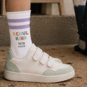 Può includere: Un calzino bianco a metà polpaccio con due strisce viola chiaro sulla parte superiore, caratterizzato dalla scritta multicolore "SCHUL KIND" e dal testo nero "ANTON" seguito dalla data "26.08.2026" sul lato, abbinato a una sneaker bianca con dettagli verde chiaro.