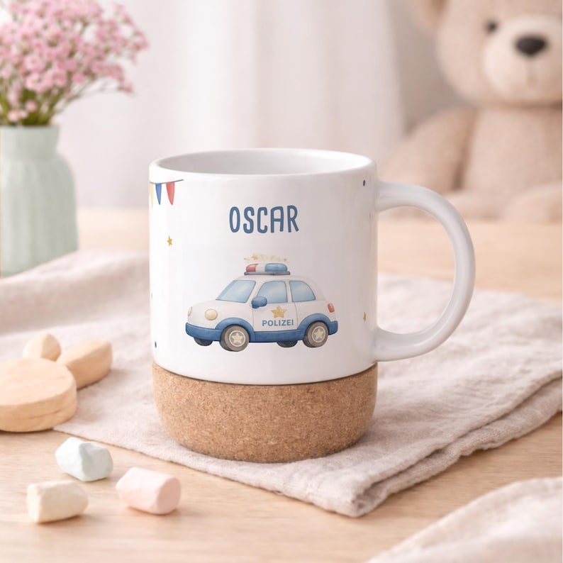 May include: White ceramic mug with a cork base, featuring the name "OSCAR" and a cartoon police car illustration. The car is white and blue with the word "Polizei" on the side. The mug has a white handle and is decorated with small stars.