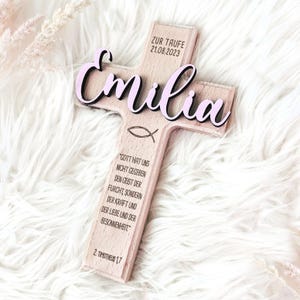 May include: A wooden cross with the name "Emilia" in pink lettering, the date "21.08.2023" and the text "Zur Taufe" on the top, and a Bible verse on the bottom.