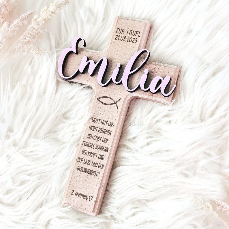 May include: A wooden cross with the name "Emilia" in pink lettering, the date "21.08.2023" and the text "Zur Taufe" on the top, and a Bible verse on the bottom.