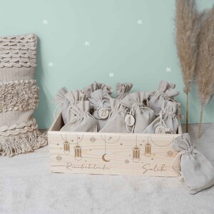 May include: A wooden box with a light wood finish and a white background. The box is filled with small burlap bags, each with a number on it. The box is decorated with a crescent moon, stars, and lanterns. The text "Ramadan Kalender" and "Salih" is engraved on the box.