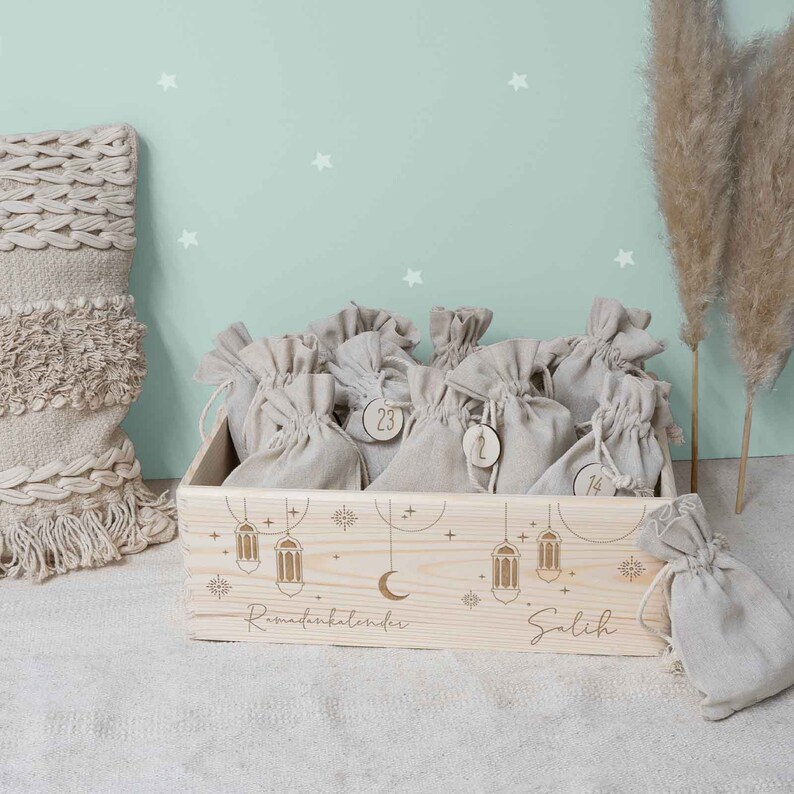 May include: A wooden box with a light wood finish and a white background. The box is filled with small burlap bags, each with a number on it. The box is decorated with a crescent moon, stars, and lanterns. The text "Ramadan Kalender" and "Salih" is engraved on the box.