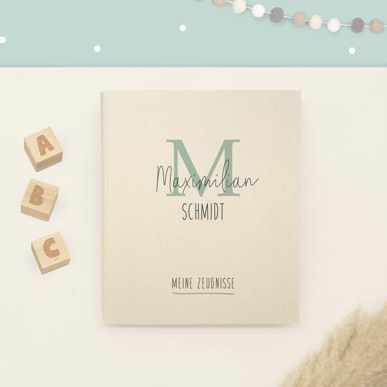 May include: A beige hardcover book featuring a large sage green letter M, the name Maximilian Schmidt, and the text MEINE ZEUGNISSE printed in black. Three wooden blocks with the letters A, B, and C are positioned to the left of the book.
