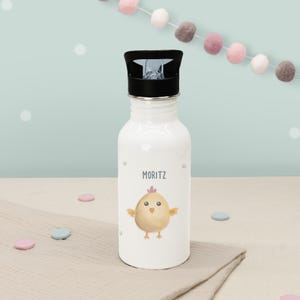 Op de afbeelding: Witte waterfles met een zwarte dop, met de naam "MORITZ" en een aquarel illustratie van een geel kuiken. De fles heeft een zilveren rand en is versierd met kleine roze stippen. Een schattig en praktisch item.