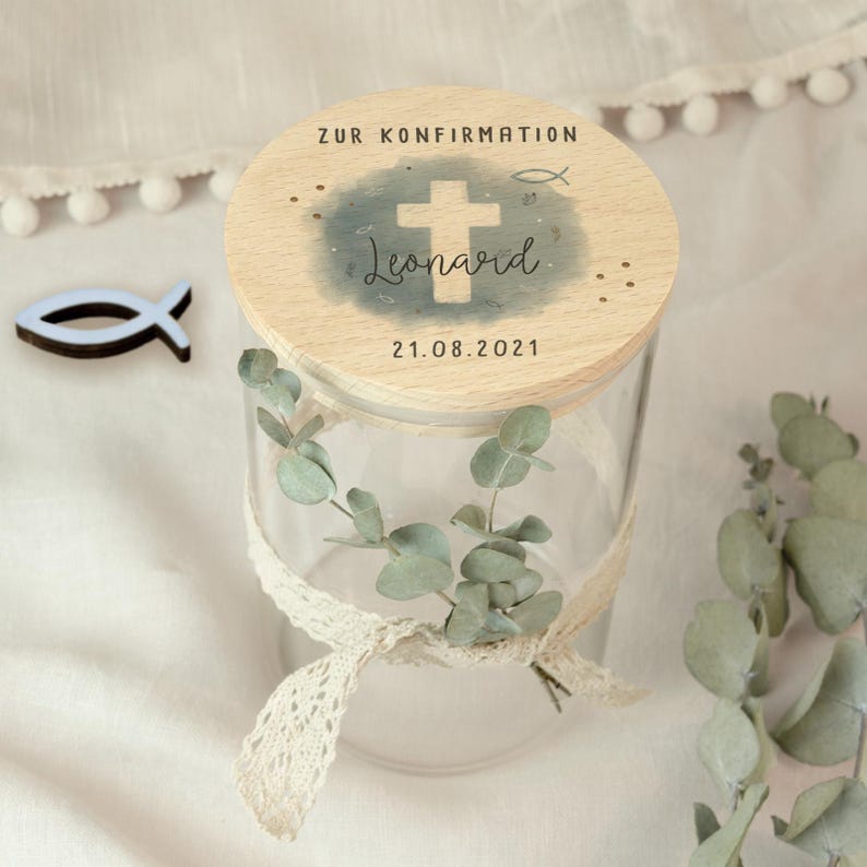 May include: A clear glass jar with a wooden lid, decorated with a cross and the name "Leonard". The lid also has the text "ZUR KONFIRMATION" and the date "21.08.2021". The jar is adorned with a lace ribbon and eucalyptus leaves.