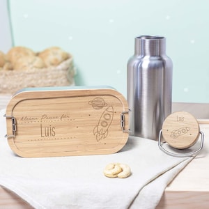 May include: A wooden lunchbox with a rocket and planet design, engraved with the text "kleine Pause f&uuml;r Luis". The lunchbox is next to a stainless steel water bottle and a matching wooden bottle stopper.