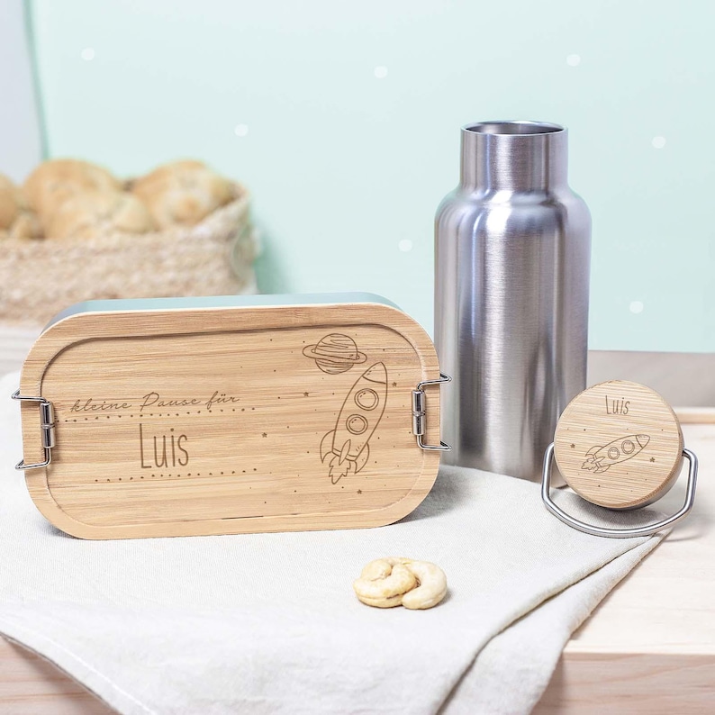 May include: A wooden lunchbox with a rocket and planet design, engraved with the text "kleine Pause f&uuml;r Luis". The lunchbox is next to a stainless steel water bottle and a matching wooden bottle stopper.
