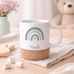 May include: White ceramic mug with a blue and gray rainbow design, the name "Noah" in a script font, and a cork base. The mug has a curved handle and is decorated with small stars.