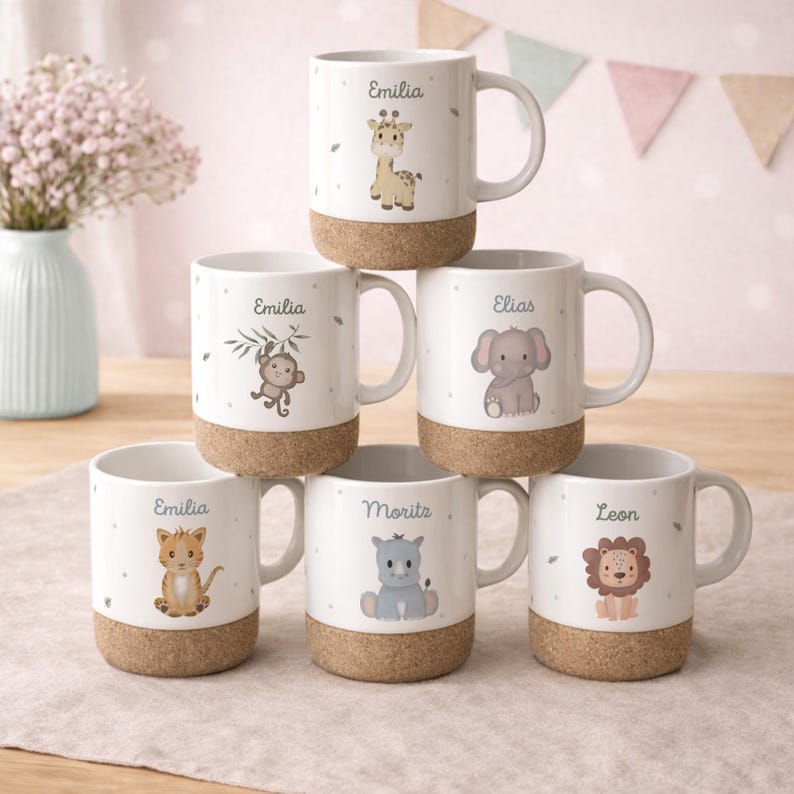 May include: A stack of white ceramic mugs with cork bases, each featuring a different cartoon animal and a name. The animals include a giraffe, monkey, elephant, cat, hippo, and lion. The names are Emilia, Elias, Moritz, and Leon.