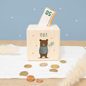 May include: A wooden piggy bank with a bear illustration and the name "THEA" on the front. A twenty euro bill is inserted into the slot on the top of the bank.