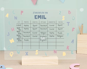 May include: A clear acrylic dry-erase board with a wooden stand. The board has a grid with the days of the week and times listed. The board says "Stundenplan von Emil" at the top. The board is surrounded by colorful letters.