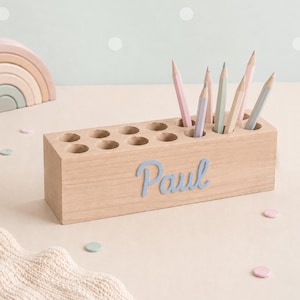 Könnte beinhalten: Ein rechteckiger Stiftehalter aus hellem Holz mit zwölf kreisförmigen Löchern, der auf der Vorderseite den Namen Paul in hellblauer Schreibschrift trägt. Mehrere Stifte in Pastellfarben stecken in den Öffnungen des Holzblocks.
