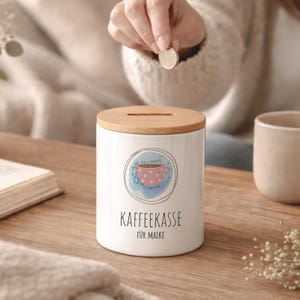Op de afbeelding: Witte keramische koffie spaarpot met een houten deksel. De spaarpot heeft een roze gestippeld koffiekopje ontwerp in een blauwe aquarel cirkel. De tekst "KAFFEEKASSE FÜR MAIKE" staat onder het ontwerp.