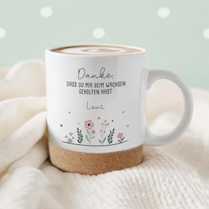 Könnte beinhalten: Weiße Keramik-Tasse mit Korkboden, gefüllt mit einem Getränk und mit einem Wirbelmuster versehen. Die Tasse zeigt Blumenillustrationen und den Text "Danke, dass du mir beim wachsen geholfen hast" und den Namen "Leve".