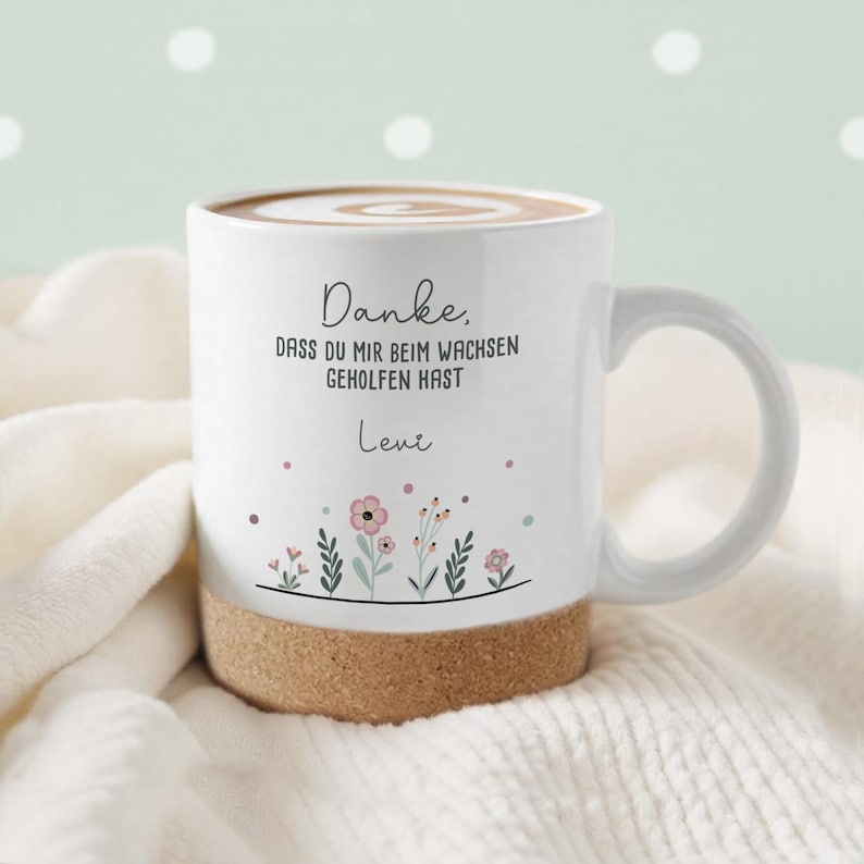 May include: White ceramic mug with a cork base, filled with a beverage and topped with a swirl design. The mug features floral illustrations and the text "Danke, dass du mir beim wachsen geholfen hast" and the name "Leve".