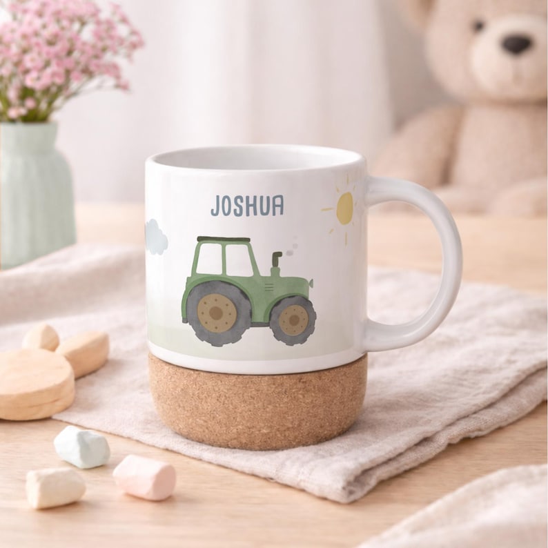 May include: White ceramic mug featuring a green tractor illustration, the name "JOSHUA" in blue, and a yellow sun. The mug sits on a cork base. A charming design for a child's drinkware.